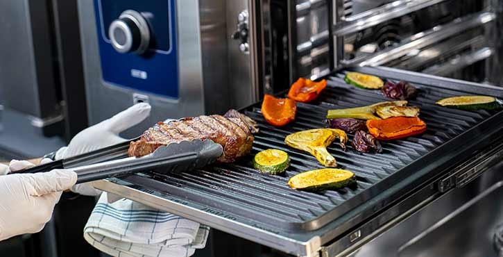 Koch, der mit einer Zange gegrilltes Steak und Gemüse aus dem iCombi Pro Ofen entnimmt.