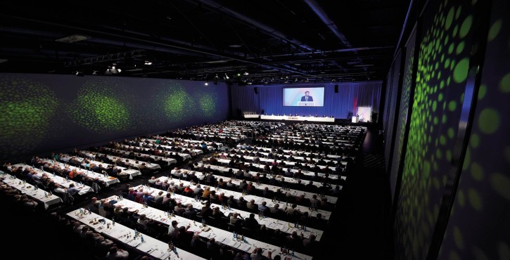Eine große Menschenmenge bei einer Versammlung in einem Konferenzsaal, mit einem Redner auf der Bühne.