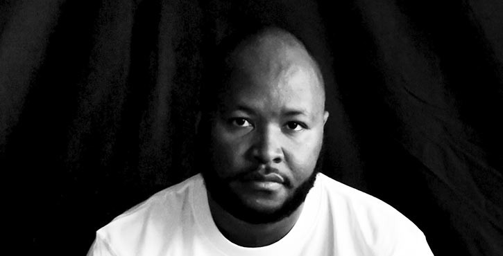 Black and white portrait of a man with a serious expression in a white T-shirt against a black background.