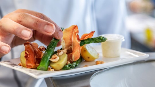 A hand garnishing a plate with asparagus, crispy prosciutto, baby potatoes, and hazelnuts, accompanied by a small container of sauce.