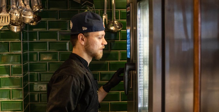 A chef wearing a cap and gloves operates a RATIONAL oven in a kitchen with green tiles and cooking tools hanging on the wall.