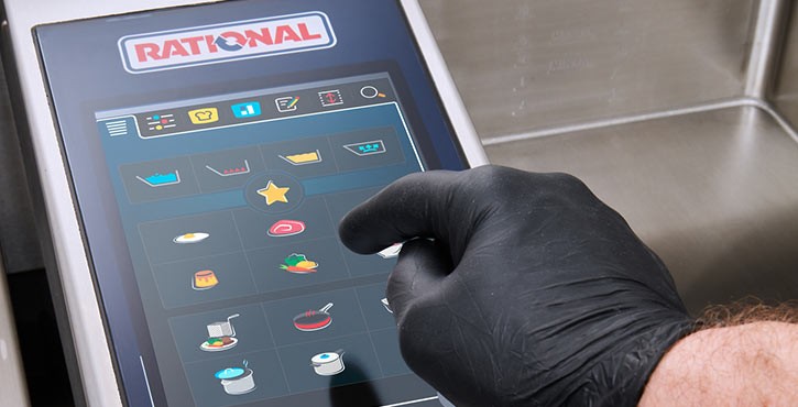 A close-up of a hand wearing a black glove interacting with the touchscreen control panel of a Rational iVario Pro appliance.