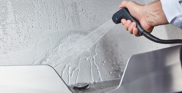A close-up of a hand holding a hand shower washing down a stainless steel surface, showcasing water droplets and steam.